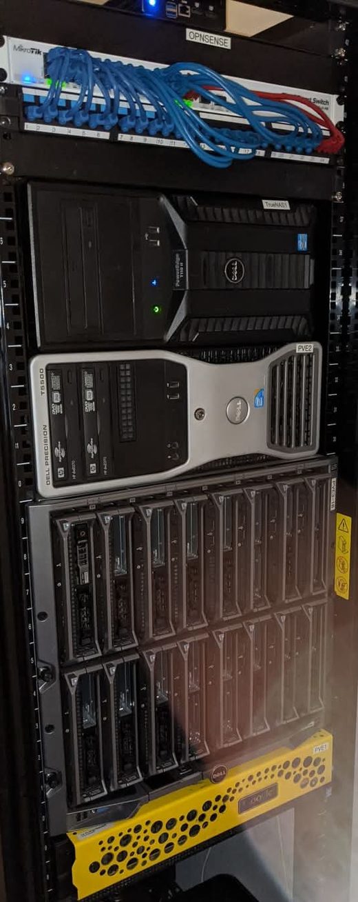 A black rack, top to bottom: a small OPNsense router above a MikroTik switch and a breakout panel thick with blue patch cables, the NAS tower labelled TrueNAS, a Dell Precision T5500 labelled PVE2, a fully populated sixteen-bay Dell M1000e blade chassis, and a yellow 1U R710 labelled PVE1 at the foot of the frame, with the UPS below it out of shot.